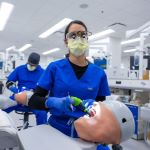 Flavia Santos Bada, a student in the advanced standing program, works on a simulation mannequin at the dental school. She wears a mask and glasses while she concentrates.