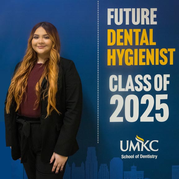dental hygiene student student wearing scrubs stands against a blue background with Future Dental Hygienist Class of 2025 and the UMKC logo behind her