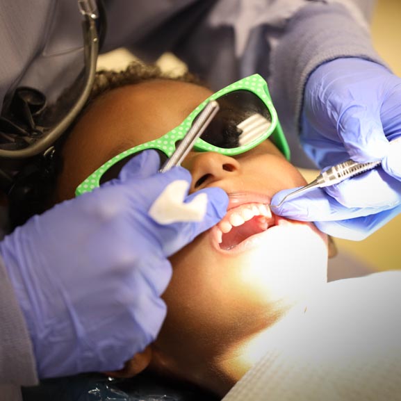 A little girl sits in a dental chair while a dental student uses instruments to examine her teeth. The girl is wearing green sunglasses.
