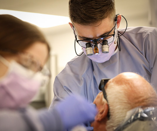 A dental student uses magnifying glasses to examine a patient in the dental chair