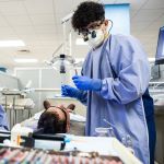 DDS student Edgardo Leiva, wearing a clinic gown and face mask, talks to a patient sitting in the dental chair.